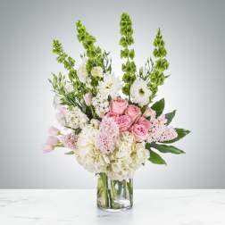 Tall arrangement of pink roses, white hydrangeas, and green bells of Ireland in a clear glass vase