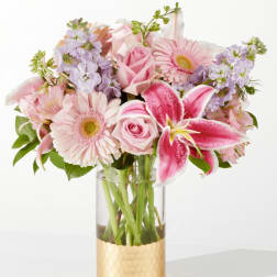Pink lilies, roses, and gerbera daisies with lavender stock in a glass vase with a gold base.