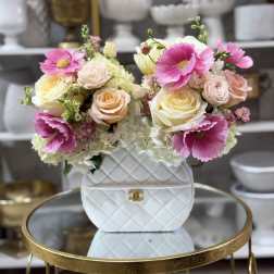 Handbag-shaped white ceramic vase filled with pink anemones, pastel roses, and white hydrangeas on a gold table