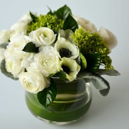 Low arrangement of white ranunculus and anemones with green hydrangea in a clear glass cylinder vase