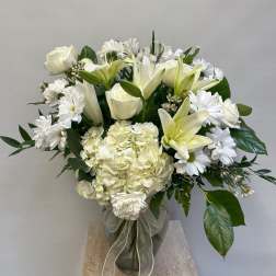 White roses, lilies, and daisies arranged in a glass vase with a ribbon.