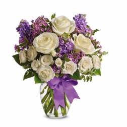 White roses and purple flowers arranged in a clear glass vase with a purple ribbon bow