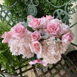 Low glass vase of light pink roses and hydrangeas arranged in a compact mound on a metal bench