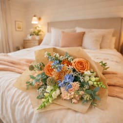 Bouquet of peach, blue, and white flowers wrapped in kraft paper on a bed