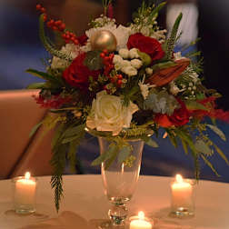 Red and white rose centerpiece with holiday greens in a clear vase, surrounded by three votive candles