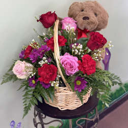 Basket arrangement of red and pink roses, red carnations, purple daisies, and a plush teddy bear.