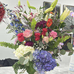 Mixed bouquet in a patterned paper cup with lilies, roses, hydrangeas, and other blooms