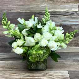 White roses and snapdragons with green accents arranged in a clear glass cube vase