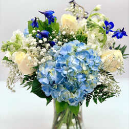 Vase arrangement with blue hydrangeas, cream roses, white blooms, and bright blue flowers