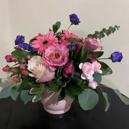 Pink and purple flower arrangement with roses and gerbera daisies in a pale pink ceramic bowl.