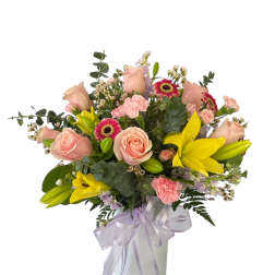 Mixed bouquet of pink roses, yellow lilies, and small blooms in a white vase with a lavender ribbon bow