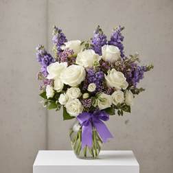White roses and purple flowers arranged in a glass vase with a purple ribbon