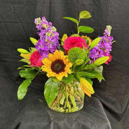 Mixed bouquet in a clear glass vase with sunflowers, pink roses, and purple blooms