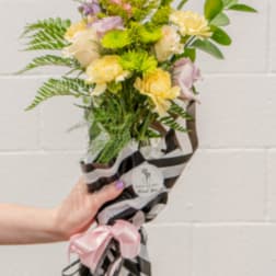Hand-tied bouquet of pink lilies, yellow carnations, and green mums in striped wrap with a pink bow