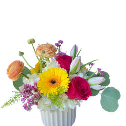 Cheerful mixed arrangement with yellow gerbera, pink roses, ranunculus and tulips in a white ceramic vase.