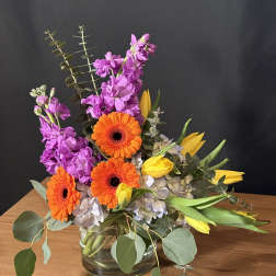 Bright mixed bouquet with orange gerbera daisies, purple flowers, and yellow tulips in a glass vase