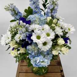 Blue hydrangeas, white daisies, and cream roses arranged in a clear glass vase.