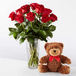 Red roses in a clear vase beside a teddy bear with a red bow.