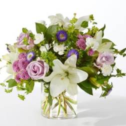 Mixed arrangement of white lilies, lavender roses, and purple asters in a clear glass vase