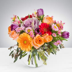 Bright mixed arrangement of orange roses, gerbera daisies and purple tulips in a round clear glass vase