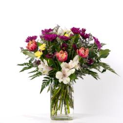 Mixed bouquet of purple daisies, pink tulips, and white blooms in a clear glass vase
