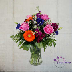 Mixed bouquet of pink roses, orange gerbera daisies, and blue blooms in a glass vase