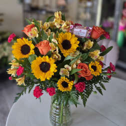 Vase arrangement of bright yellow sunflowers, orange roses, pink carnations, and yellow alstroemeria.