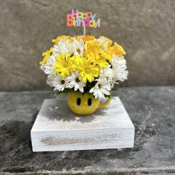 Yellow and white daisy bouquet in a smiley-face mug with a Happy Birthday topper