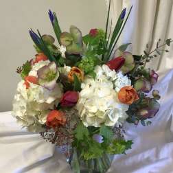 Mixed bouquet of white hydrangeas, orange blooms, and purple flowers in a glass vase