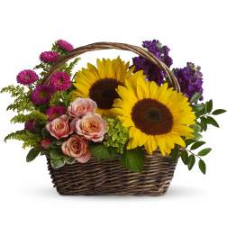 Wicker basket arrangement with bright yellow sunflowers, pink roses, purple blooms, and accent flowers.
