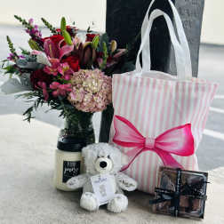 Bouquet with pink and red flowers beside a striped gift bag, candle, teddy bear, and chocolates