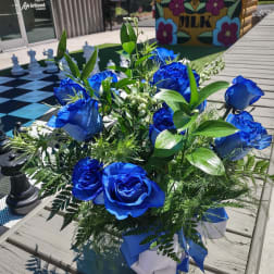 Blue roses arranged with greenery in a white container on a boardwalk