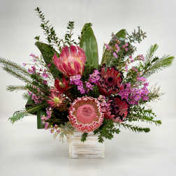 Arrangement of pink protea and small magenta blooms in a rustic whitewashed cube vase