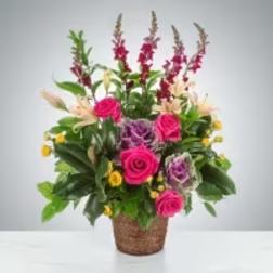 Tall mixed arrangement of pink roses, snapdragons, and purple blooms with yellow accents in a woven basket container