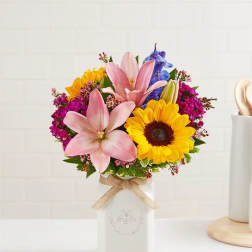 Colorful bouquet of pink lilies, sunflowers and purple blooms in a white ceramic jar with twine bow.