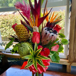 Tall tropical arrangement with red and pink flowers and protea in a clear glass vase with a red bow.