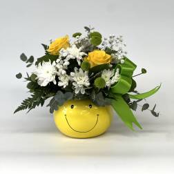 Yellow and white flower arrangement in a smiley-face pot with a green ribbon