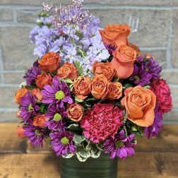 Bright arrangement of orange roses, purple daisies, and pink carnations in a green cube vase with Happy Birthday pick