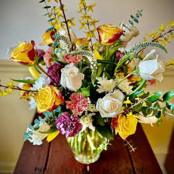 Mixed arrangement of yellow and white roses with pink carnations in a clear glass vase on a wooden table.