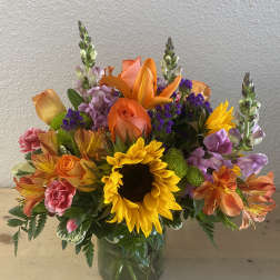 Colorful bouquet with sunflowers, roses, lilies, and purple filler in a glass vase