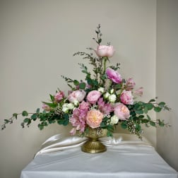 Asymmetrical arrangement of pink and peach roses and ranunculus in a gold pedestal vase with trailing greenery.