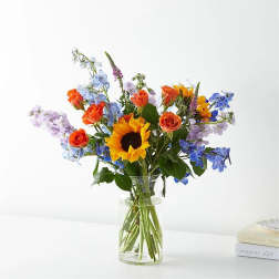 Mixed bouquet with a sunflower, orange roses, and blue blooms in a clear glass vase