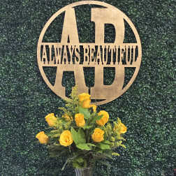 Yellow roses in a clear glass vase with a gold monogram sign behind them