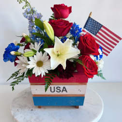 Red roses and white lilies in a patriotic wooden container with a small U.S. flag.