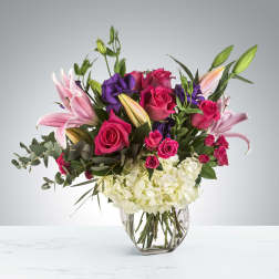 Mixed arrangement of hot pink roses, pink lilies, purple blooms, and white hydrangea in a clear glass vase