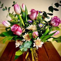 Pink roses, tulips, and pale peach mums with purple accents in an amber glass vase.
