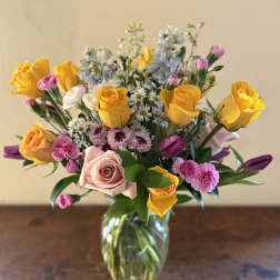 Mixed bouquet of yellow roses, pink roses, purple tulips, and small white daisies in a clear glass vase