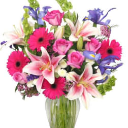 Mixed bouquet of pink gerberas, roses, lilies, and purple flowers in a glass vase