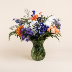 Mixed bouquet of orange, white, blue, and purple flowers in a green glass vase