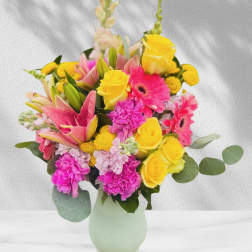 Bright mixed bouquet of yellow roses, pink lilies, and gerbera daisies in a pale green vase